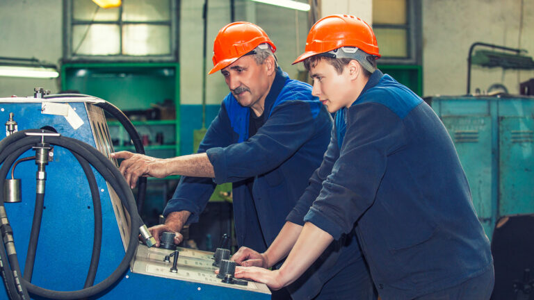 An aged worker trains a younger worker in front of an equipment